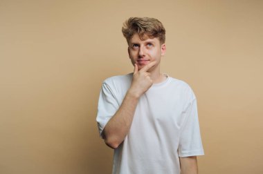 Açık kahverengi saçlı genç bir adam düşünceli bir şekilde çenesini elinde dinlendiriyor, hafifçe yukarı bakıyor. Yumuşak bir bej arkaplan önünde meraklı ve dalgın görünüyor..