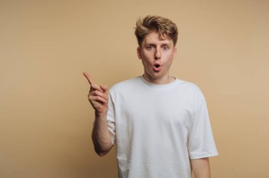 Saçları dağınık genç bir adam heyecan ifade ediyor, parmak kaldırırken düz bir arkaplan karşısında şaşırmış bir yüz ifadesi gösteriyor. Beyaz gömleği yumuşak renkle çelişiyor..