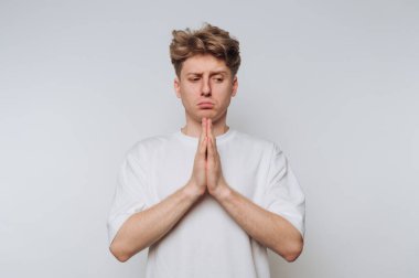 Genç bir adam ciddi bir ifadeyle ayakta duruyor, elleri önünde sıkıca bağlı. Minimalist, aydınlık bir ortamda derin düşüncelere dalmış ya da endişeli görünüyor..