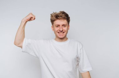 Kıvırcık saçlı genç bir adam zafer jesti olarak kolunu esnerken geniş bir şekilde gülümsüyor. Düz beyaz bir tişört giymiş, arka planı aydınlık, pozitiflik ve coşku yayıyor..