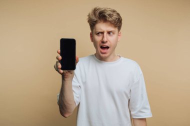 Genç bir adam, tarafsız bir arkaplan önünde akıllı telefonu tutarken şok geçirir. Yüzündeki ifade, bu iç mekan sahnesinde şaşkınlık ve heyecan yaratıyor..