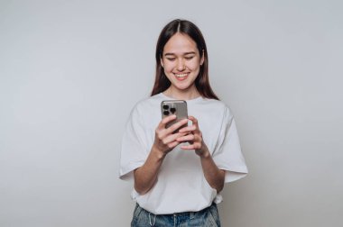 Genç bir kadın kapalı alanda parlak, minimalist bir alanda duruyor, akıllı telefonuyla konuşurken gülümsüyor. Günlük kıyafetleri basit bir arkaplanı tamamlıyor..