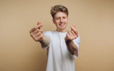 Genç bir adam, kollarını ileri uzatırken parlak bir şekilde gülümseyen tarafsız bir arkaplanda duruyor. Neşeli el hareketleri yapar, dostluk ve neşe iletir..