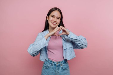 Genç bir kadın elleriyle bir kalp şekli oluşturarak mutluluğunu ifade eder. Kendine güveni tam pembe bir arka planda duruyor, sıradan bir tarz sergiliyor.