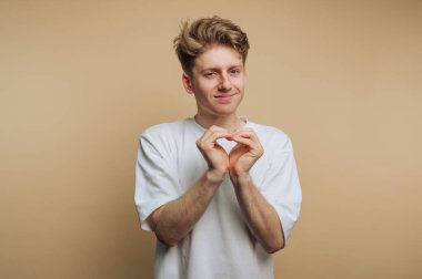 Basit beyaz tişört giyen genç bir adam elleriyle kalp şeklini oluşturarak sıcaklığını ifade eder. Tarafsız bir arkaplanda dostane bir gülümsemesi var, pozitif enerji ve şefkat saçıyor..