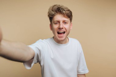 Açık sarı saçlı neşeli bir genç adam coşkuyla gülümsüyor ve koluyla elini uzatıyor. Düz beyaz bir tişört giyiyor ve açık bej bir arka planda mutlu görünüyor..