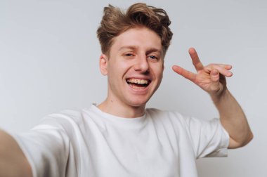 Genç bir adam sade bir arka plana karşı durur, selfie çekerken neşeyle ışıldar. Tek eliyle barış işareti yapıyor, cana yakın ve neşeli görünüyor..