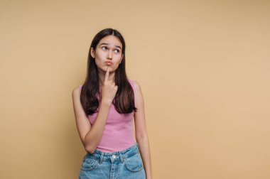 Uzun siyah saçlı genç bir kadın, parmağını çenesinde tutarak düşünceli bir şekilde ayakta duruyor. Pembe bir kolsuz bluz ve kot şort giyiyor. Sarı bir arka plan, anı güzelleştiriyor..