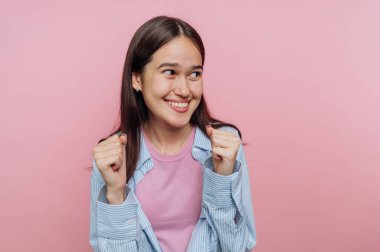 Genç bir kadın, elleri havada, rahat bir kıyafetle gülümseyen bir şekilde sevinç ifade eder. Yumuşak pembe arka plan onun neşeli tavrını güçlendiriyor ve canlı bir atmosfer yaratıyor..