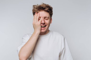 Açık renkli bir gömlek giyen genç bir adam stüdyoda duruyor, bir gözünü neşeyle kapatıp neşeyle gülümsüyor. Arka plan tarafsız ve ifadesini vurguluyor..