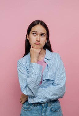 Genç bir kadın düşünceli bir ifadeyle ayakta duruyor, elleri çenesinde, dik dik bakıyor. Uzun koyu renk saçları var ve rahat bir bluzun üzerine çizgili bir gömlek giyiyor, canlı pembe bir arka fona kurulmuş..