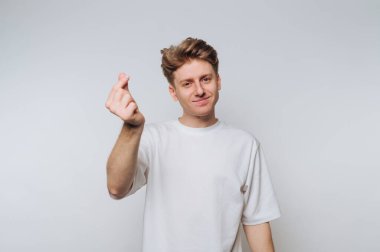 Tarzlı saçları olan genç bir adam güven içinde duruyor, sevgi ve takdiri ifade eden bir el hareketi yapıyor. Sıradan beyaz bir tişört giyiyor ve yüzünde hafif bir gülümseme var..