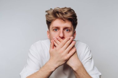 Açık kahverengi saçlı genç bir adam ağzını iki eliyle kapayarak sürpriz bir yüz ifadesi sergiliyor. Arka plan yumuşak, nötr bir gölge, duygularını vurguluyor..