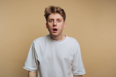 Sarı saçlı genç bir adam, bronzlaşmış bir geçmişe karşı şok ve inançsızlık ifadesi sergiliyor. Geniş gözleri ve ağzı beklenmedik bir tepki veriyor..