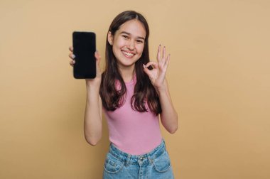 Genç bir kadın açık renkli bir duvarın karşısında duruyor, bir elinde akıllı bir telefon tutarken geniş bir gülümsemeyle. Diğer eliyle iyi bir jest yapıyor, pozitiflik yayıyor..