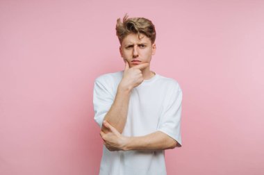 Genç bir adam pembe bir arka fonun önünde duruyor, eli çenesinde düşünceli görünüyor. Düz beyaz bir tişört giyiyor ve sanki derin düşüncelere dalmış gibi biraz şaşkın bir ifadesi var..