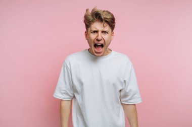 Saçları dağınık genç bir adam hayal kırıklığı içinde bağırıyor, stüdyo seansı sırasında açık pembe bir arkaplan önünde ham duygular sergiliyor. İfadesi yoğunluğu iletir..