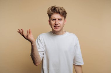 Genç adam bej bir arka plana karşı duruyor, kafası karışmış görünüyor, kafası karışmış ve hayal kırıklığı yaşıyor. Onun günlük kıyafetleri rahat atmosfere katkı sağlar..