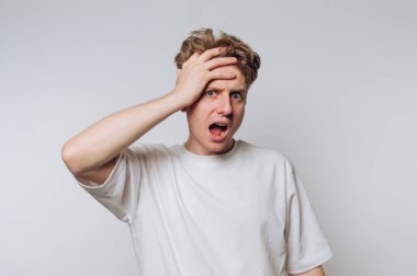 Genç bir adam, tek eliyle kafasını tutarak, bir şaşkınlık veya hayal kırıklığı ifadesi göstererek sade bir arkaplanda duruyor. Onun günlük beyaz gömleği onun duygusal yüzüyle çelişiyor..