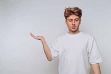 Genç bir adam minimalist bir çevrede duruyor, tek eliyle hareket ederken düşünceli bir yüz ifadesi sergiliyor, bir sorgulama ya da açıklama hissi veriyor..