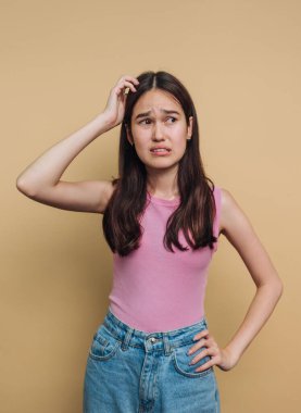 Genç bir kadın, elleri başının üzerinde, pembe bir atlet ve mavi kot pantolon giyerek kafası karışmış bir ifade sergiliyor. Düz bir zemine karşı duruyor, belirsizlik hissi veriyor..