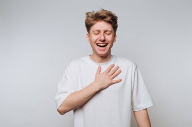 Genç bir adam mutluluğu samimi bir kahkaha ile ifade eder, gözleri kapalı ve eli göğsünde, gerçek bir sevinç anı iletir. Basit, hafif bir arkaplana karşı duruyor..