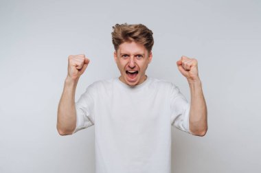 Genç bir adam zaferle yumruklarını kaldırarak sevinç ve heyecan ifade eder. Onun enerjik duruşu ve geniş gülümsemesi parlak, minimalist bir stüdyoda bir başarı duygusu taşır..