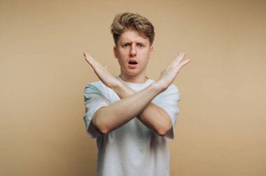 Kollarını vücudunun önünde kavuşturmuş, şaşırmış bir ifadeyle duran genç bir adam. Bir şeyi reddediyor ya da reddediyor gibi görünüyor. Arka plan yumuşak nötr bir renktir..