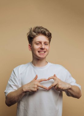Beyaz tişörtlü genç bir adam elleriyle kalp şekli yapıyor, özgüvenle gülümsüyor. Tarafsız arka plan onun neşeli ifadesini ve neşeli enerjisini vurguluyor..