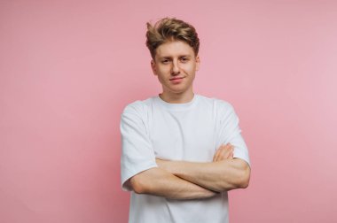 Kolları çapraz duran genç bir adam rahat bir pozda, parlak pembe bir arka fonun önünde, beyaz bir tişört giyiyor. İfadesi güven ve ulaşılabilirlik taşıyor..