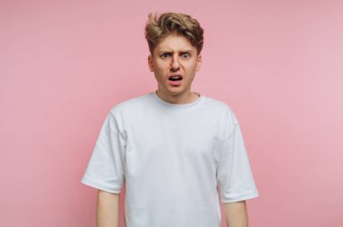 Genç bir adam pembe bir arka plana karşı karışık bir ifadeyle duruyor, basit beyaz bir tişört giyiyor. Gözleri şaşkınlık içinde, beklenmedik bir anı yakalıyor..