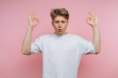 Genç bir adam meditasyon pozu veriyor, elleriyle 'OK' jesti yaparken yumuşak pembe bir arka plana sükunet yayıyor, farkındalığı ve rahatlamayı teşvik ediyor..
