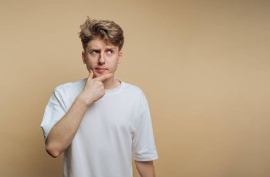 Açık kahverengi saçlı genç bir adam, eli çenesinde, bej bir arka planda düşünceli bir şekilde poz verir. İfadesi bir konu hakkında düşünmeyi ya da merakı akla getiriyor..