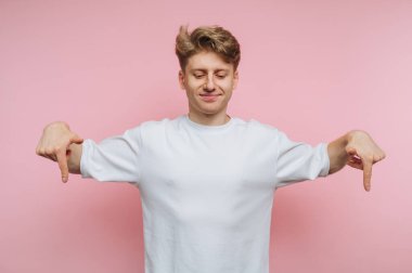 Genç bir adam yumuşak pembe bir arka fonun önünde duruyor. O, iki eliyle neşeyle aşağıya işaret ederken geniş bir şekilde gülümser. Gündelik kıyafetleri neşeli havayı tamamlıyor..