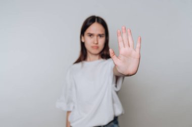 Genç bir kadın düz bir arkaplana karşı duruyor ve dur işareti olarak elini kaldırıyor. İddialı görünüyor, güçlü bir mesaj ya da duygu iletiyor. Işık yumuşak ve nötr..