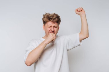 Saçları dağınık genç bir adam geniş bir alana yayılmış, yüzü bitkinlik belirtileri gösteriyor. Düz bir arkaplan karşısında duruyor, bir kolunu bir can sıkıntısı jesti olarak kaldırıyor..