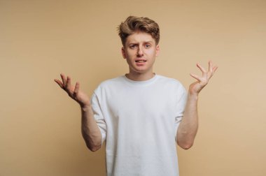 Genç bir adam sorgu jestinde ellerini kaldırarak kafa karışıklığını ifade eder. Kısa, açık renkli saçları var ve tarafsız bir arkaplana karşı düz beyaz bir gömlek giyiyor..