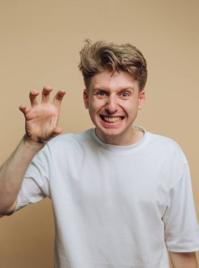 Sarışın kıvırcık saçlı genç bir adam, el pençe hareketini taklit ederken geniş bir gülümseme sergiliyor. Tarafsız arka plan onun neşeli ifadesini vurguluyor..