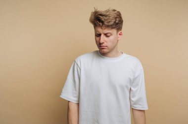 Açık kahverengi saçlı genç bir adam düz bir bej arka planda duruyor. Sıradan beyaz bir tişört giyiyor ve aşağıya bakıyor, derin düşüncelere daldığını gösteriyor..