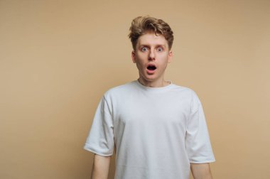Genç bir adam geniş gözleri ve ağzı açık bir şekilde ayakta duruyor ve şok olduğunu ifade ediyor. Düz beyaz bir tişört giyiyor ve arka planı yumuşak bir bej. Bu da şaşkınlığının dramatik etkisini artırıyor..
