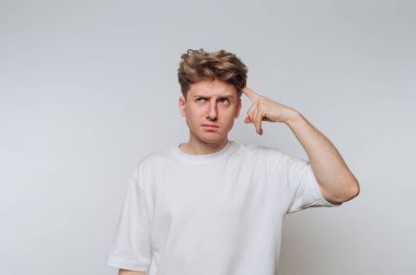 Genç bir insan, düşünceli bir ifadeyle beyaz bir zemine karşı duruyor. Alnına sanki kafa karıştırıcı bir soru hakkında derin düşüncelere dalmış gibi dokunuyor..