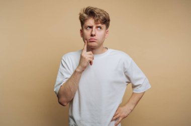 Genç bir adam, beyaz gömlekli, bej bir arka plana karşı düşünceli bir şekilde ayakta duruyor. Parmağı çenesinde duruyor. Düşüncelerini derin derin düşünüyor..