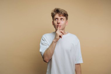 Tarafsız bir arkaplanda duran genç bir adam, düz beyaz bir tişört giyiyor ve işaret parmağını dudaklarına dayıyor, sessizlik ya da gizlilik ihtiyacını öne sürüyor..