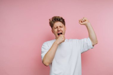 Genç bir adam, sıradan beyaz bir gömlek giyerken gözleri kapalı ve kollarını uzatarak esniyor. Pembe arkaplan o ana canlı bir atmosfer katıyor..