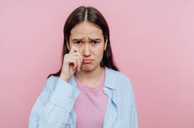Genç bir kadın pembe bir arka planın önünde duruyor, ifadesi üzüntüyü yansıtıyor. Gözlerinden bir damla gözyaşı siler, düşüncelere daldığı duygusal bir anı somutlaştırır..