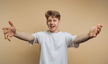 Neşeli genç bir adam, sanki birini karşılıyormuş gibi kolları açık, yumuşak bir stüdyoda duruyor. Neşeli ifadesi pozitiflik yayıyor, sıcak bir atmosfer yaratıyor..