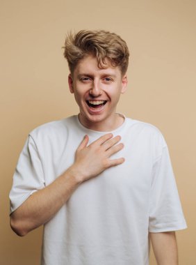 Kıvırcık saçlı genç bir adam neşeli bir gülümseme sergiliyor, yürekten gülüyor. Bir eli göğsünde, bej bir zemin üzerine sıcaklık ve neşe saçıyor..