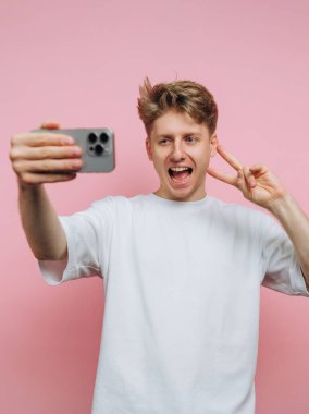 Genç bir adam gülümsüyor ve selfie çekerken barış işaretiyle poz veriyor. Yumuşak pembe bir arka plana karşı duruyor, akıllı telefonuyla eğlenceli bir anı yakalıyor..