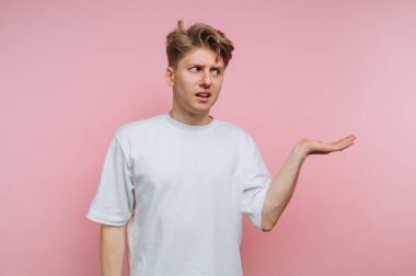 Saçları dağınık genç bir adam pembe bir duvarın önünde duruyor, elleriyle hareket ederken şaşkın bir yüz ifadesi sergiliyor. Düz beyaz bir tişört giyiyor..