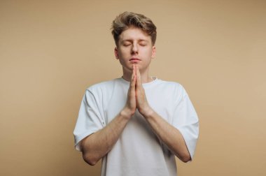 Genç bir adam elleri ve gözleri kapalı dua pozisyonunda duruyor. Odaklanmış ve huzurlu görünüyor. Konsantrasyonu ve iç huzuru simgeliyor..